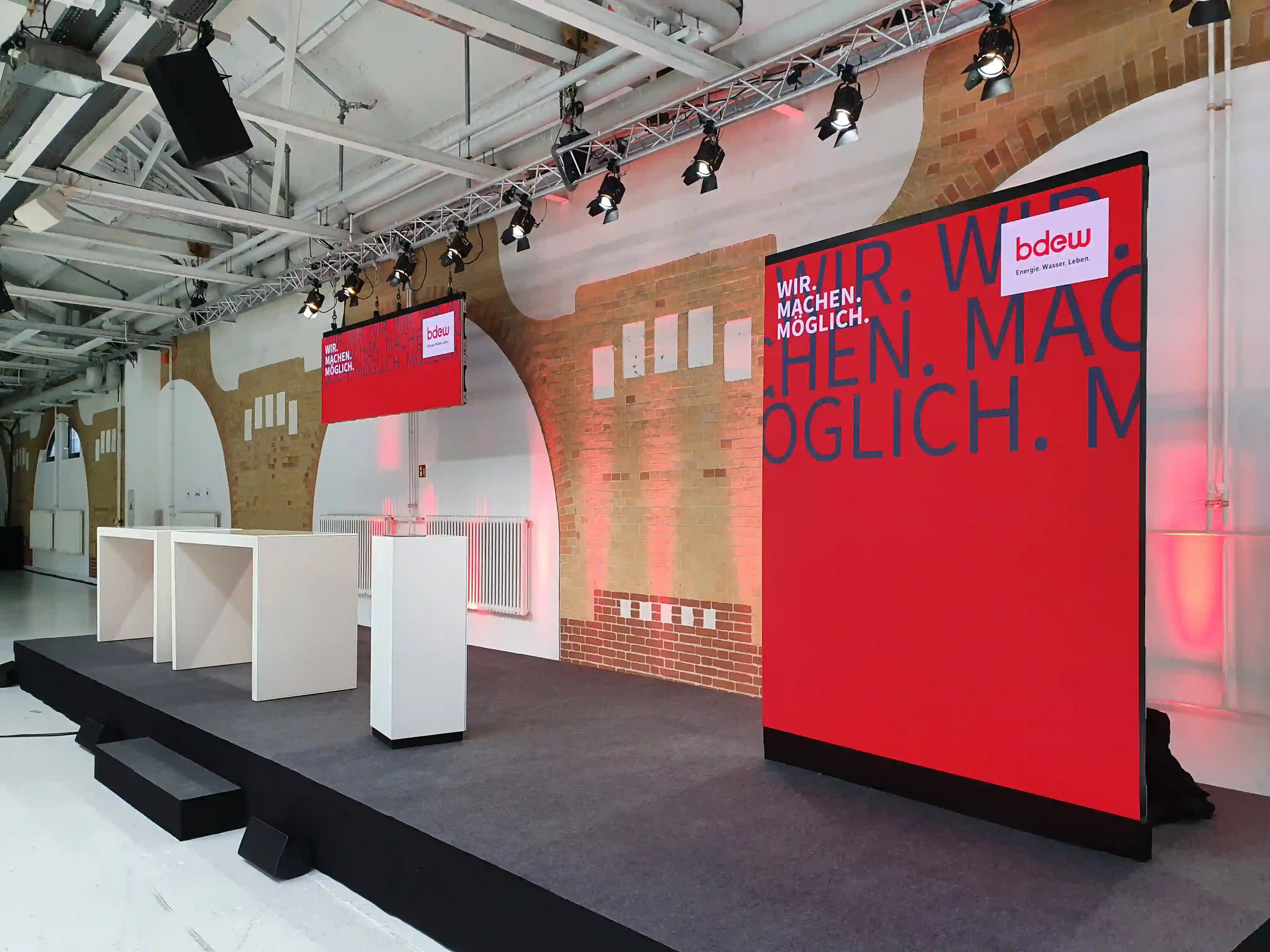 Messestand mit großem rotem Display und bdew-Logo, weißen Elementen und Ziegelmauer-Hintergrund in Industriehalle