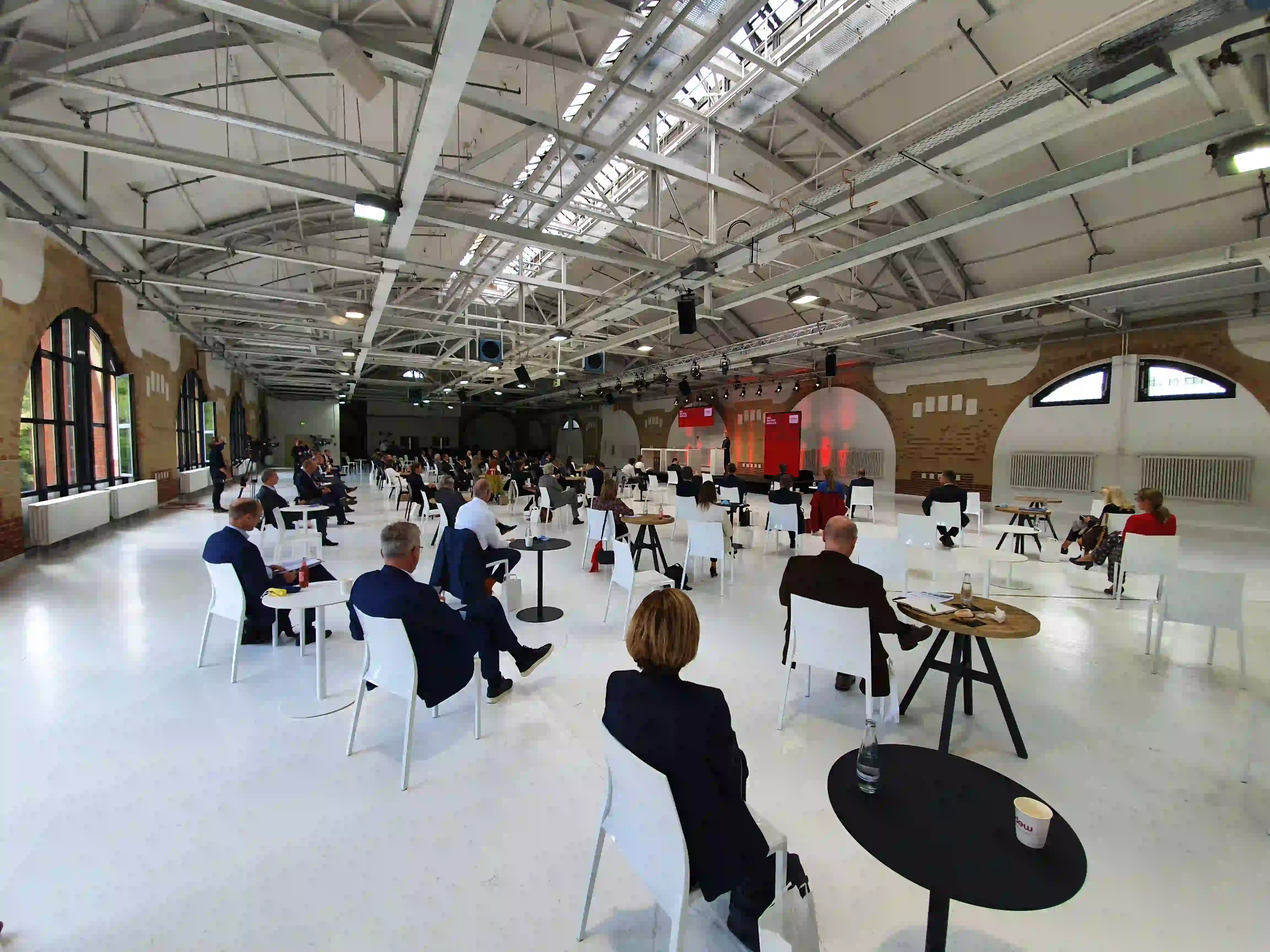 Konferenzteilnehmer sitzen mit Abstand an Tischen in großer Industriehalle mit Stahlkonstruktion und roten...