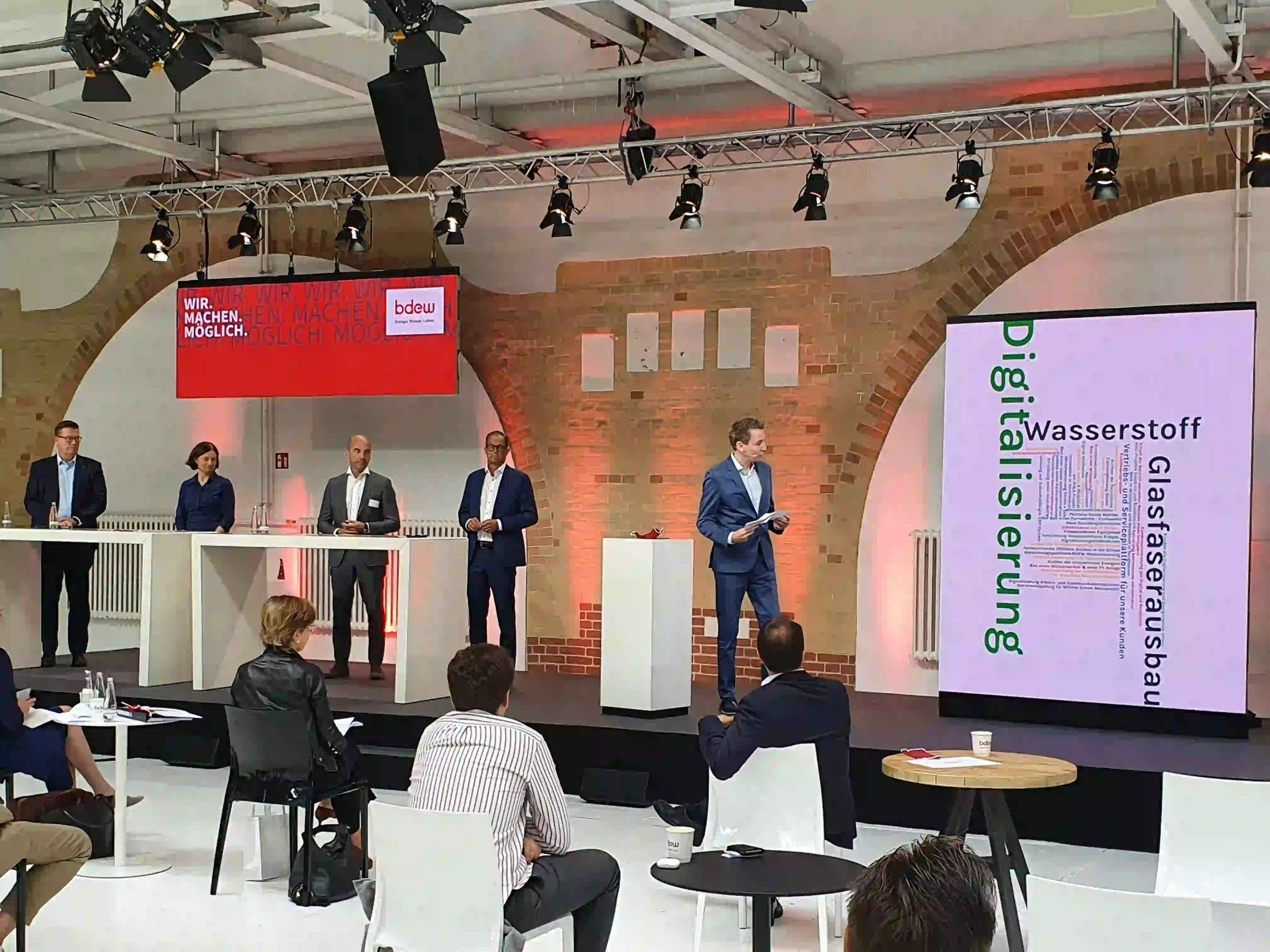 Konferenz-Bühne mit fünf Sprechern, Publikum im Vordergrund, rote Beleuchtung, BDEW-Banner und Großbildschirm zum...