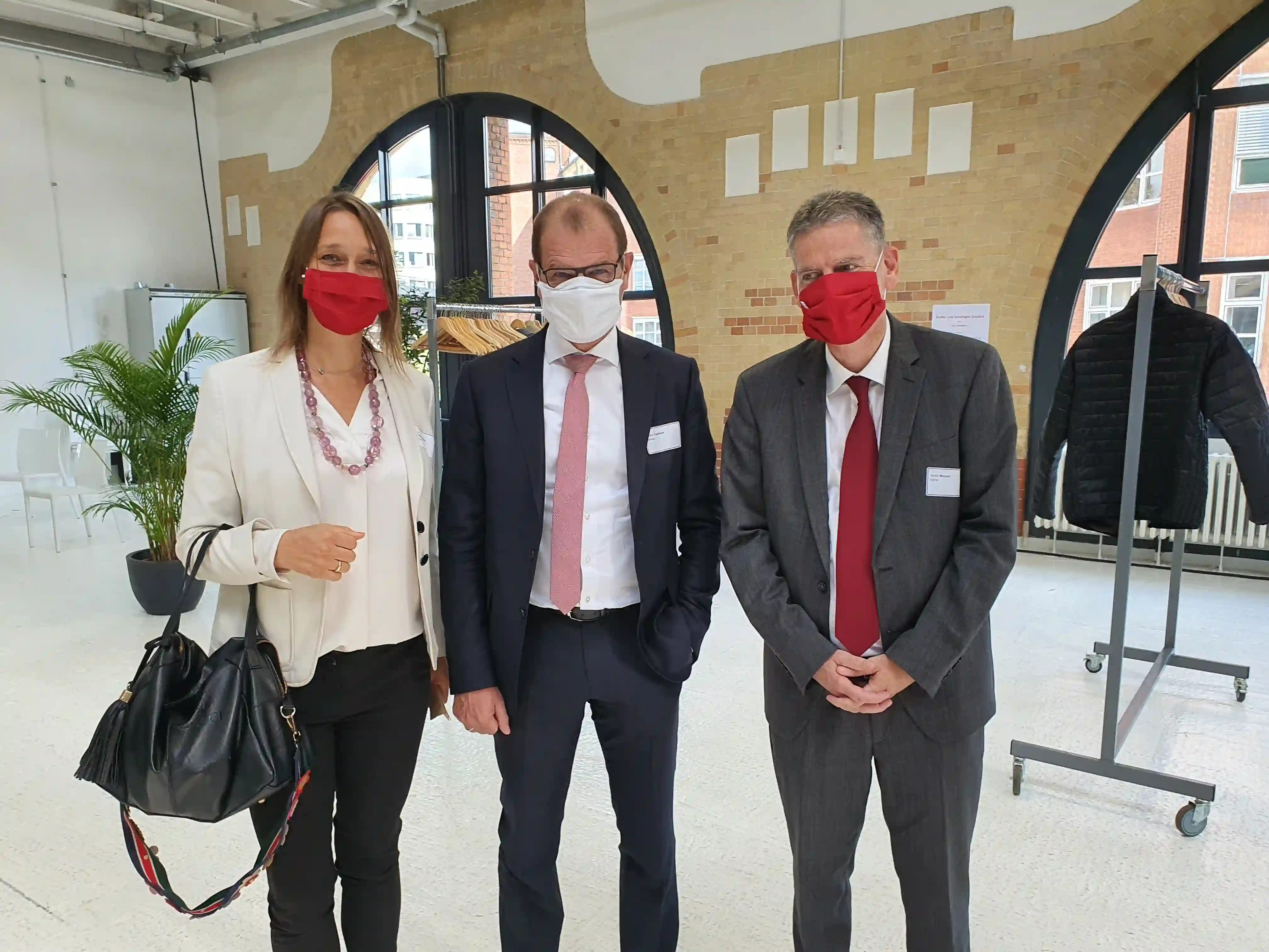 Vier Personen in Business-Kleidung mit Mund-Nasen-Masken stehen in modernem Büroraum mit Backsteinwand