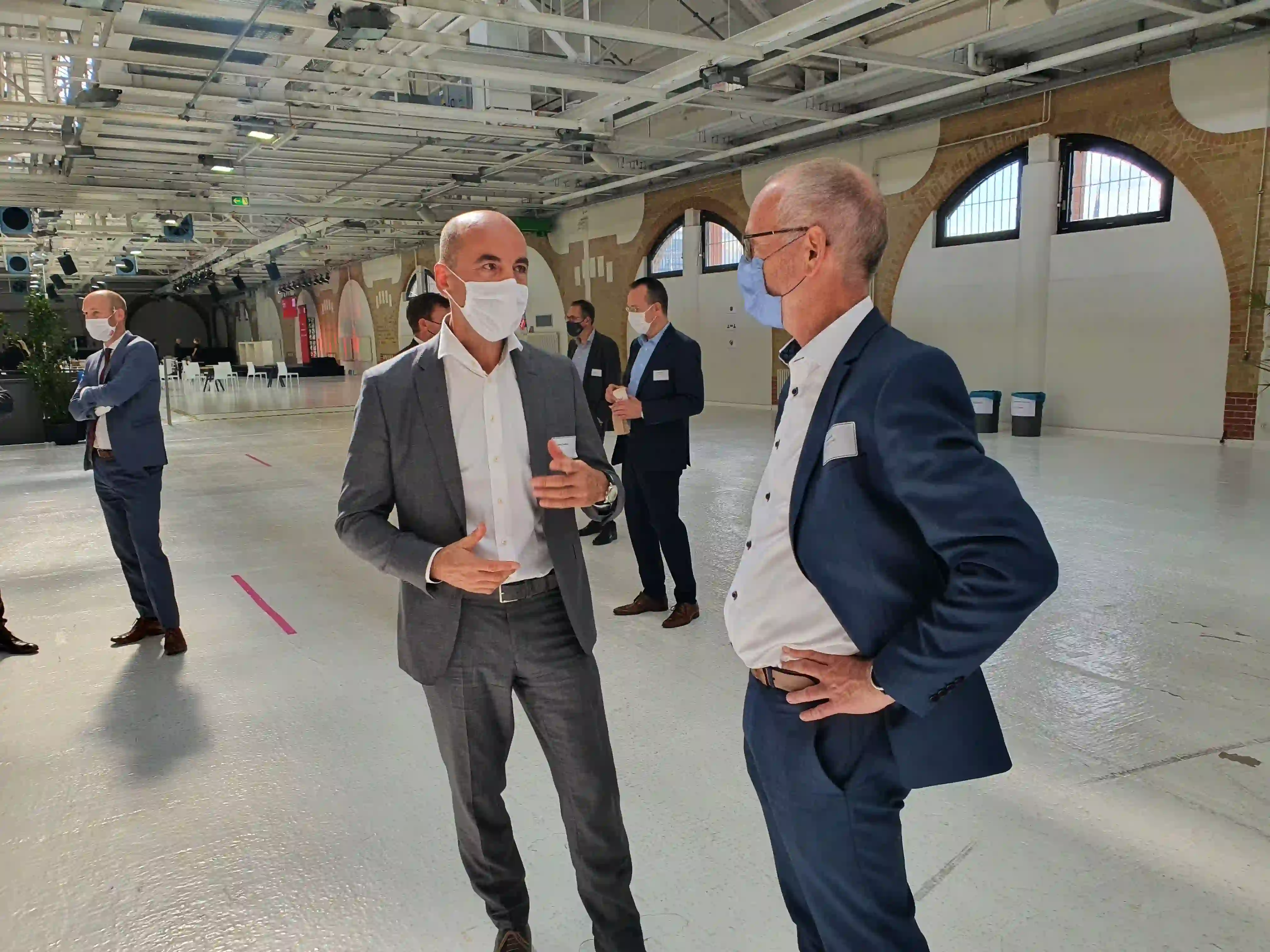 Mehrere Personen in Anzügen und Masken stehen sich in einer leeren Industriehalle gegenüber und unterhalten sich.