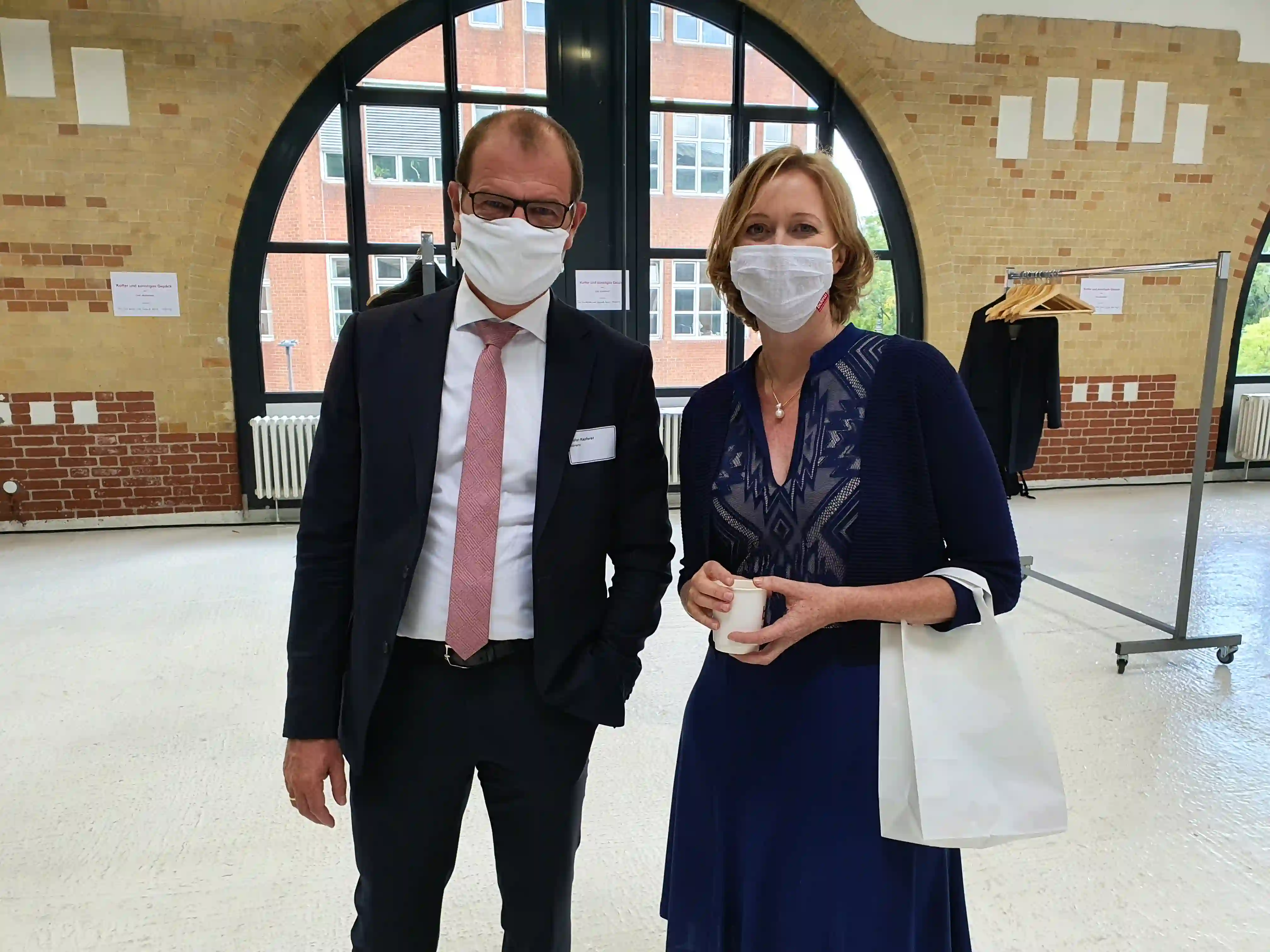Zwei Personen mit Schutzmasken in einem Gebäude mit Backsteinwand und großem Rundfenster stehen sich gegenüber.