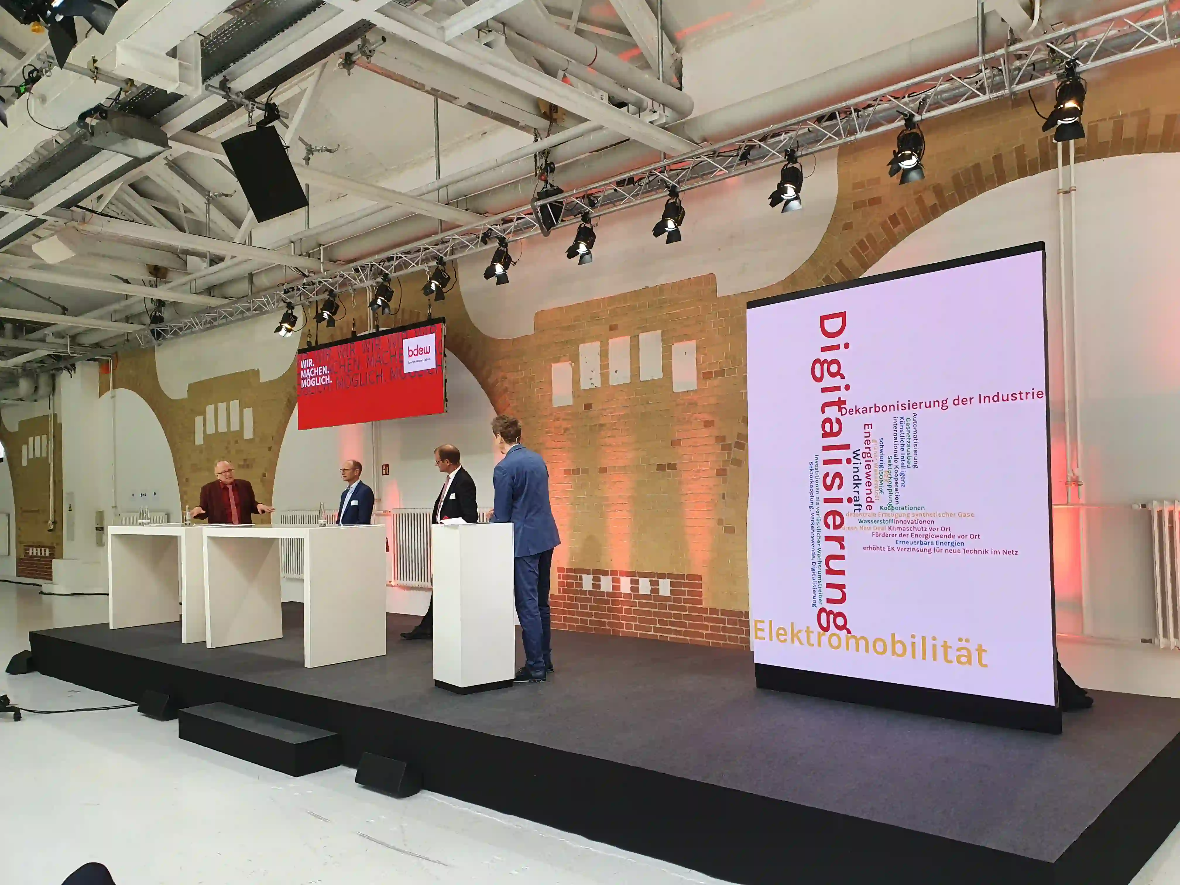 Podiumsdiskussion auf Bühne mit vier Personen, großem Display zeigt Zahlen zu Milliarden EUR, Zuschauerplätze im...