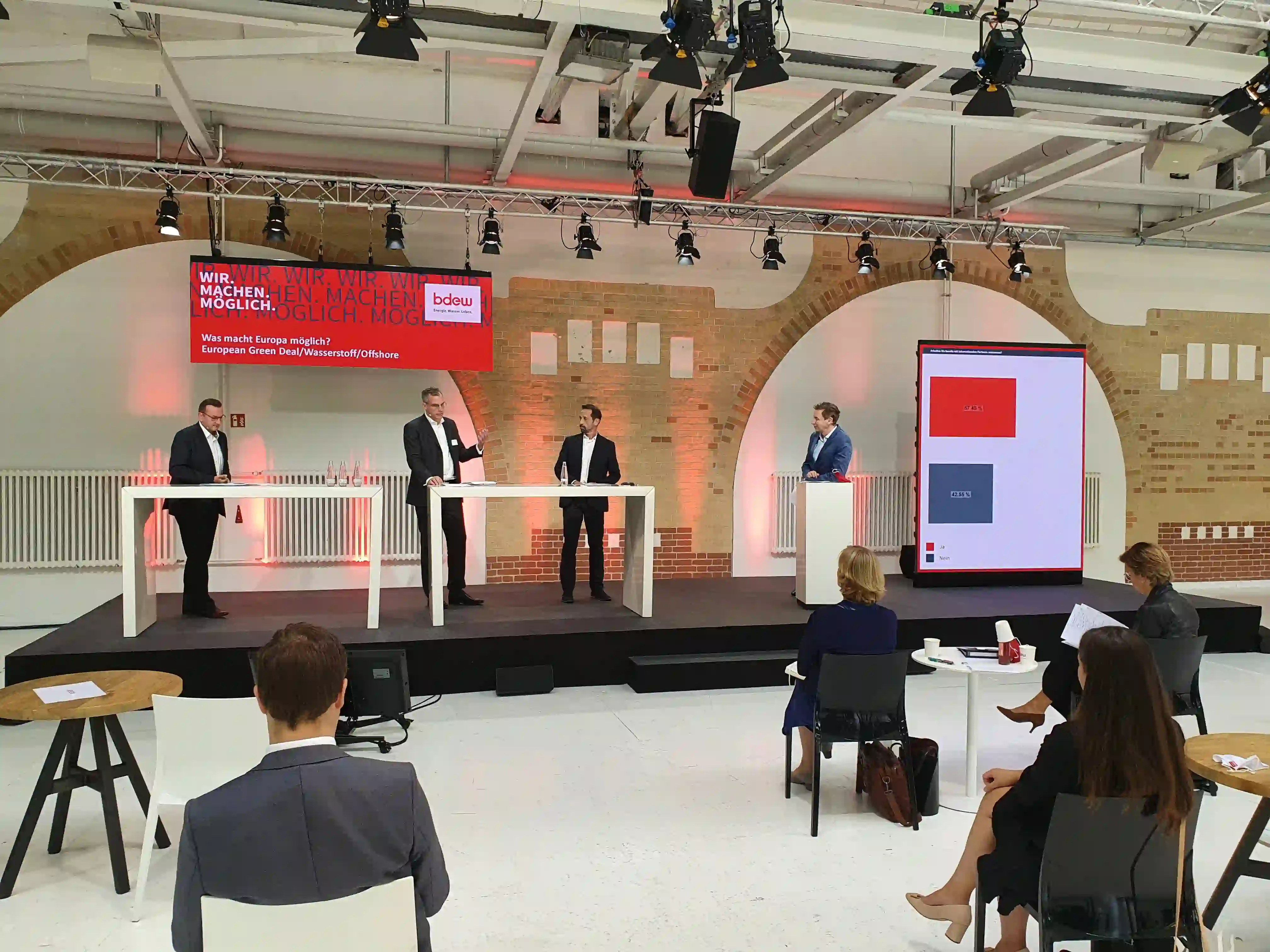 Podiumsdiskussion mit vier Männern an weißen Stehtischen auf schwarzer Bühne, rotem Boden, BDEW-Banner und Display...