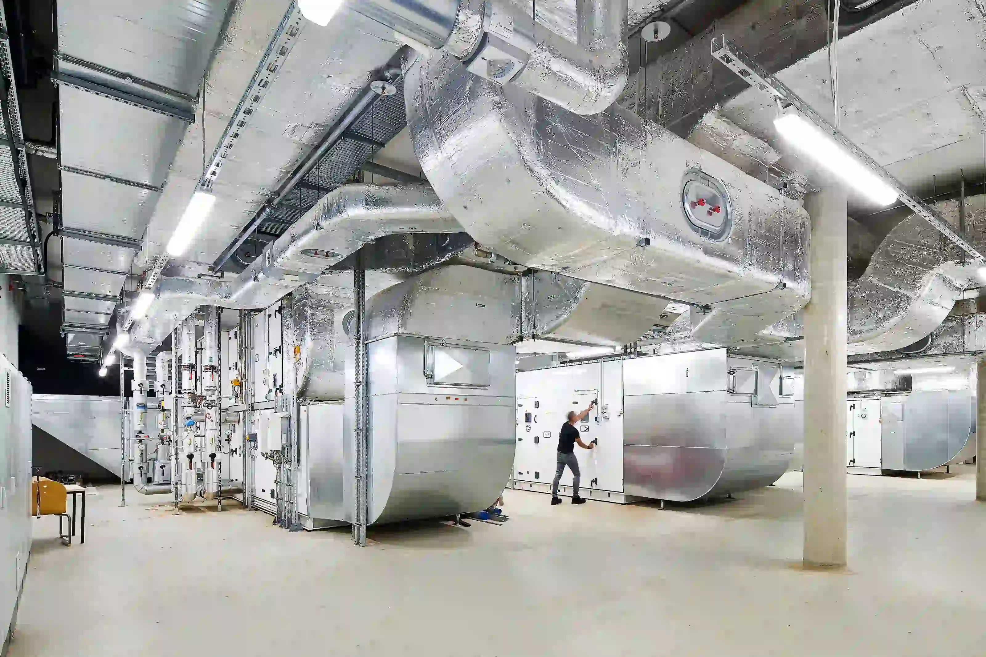 Techniker arbeitet in Maschinenhalle mit großen silbernen Wärmeanlagen, Rohrleitungen und Steuergeräten an der Decke