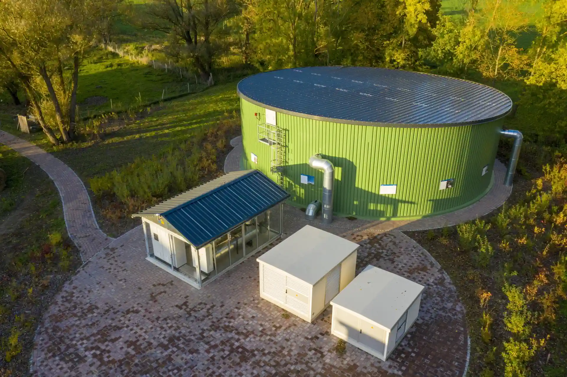 Luftaufnahme: Grüner zylindrischer Wärmespeicher mit Solardach, daneben kleinere Technikgebäude, umgeben von...