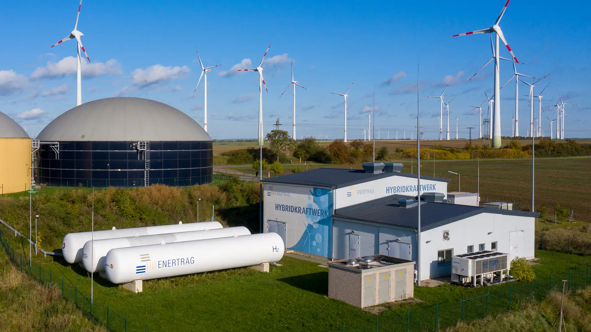 Wasserstoff-Elektrolyseanlage mit Speichertanks vor Windkraftanlage in Flachlandschaft