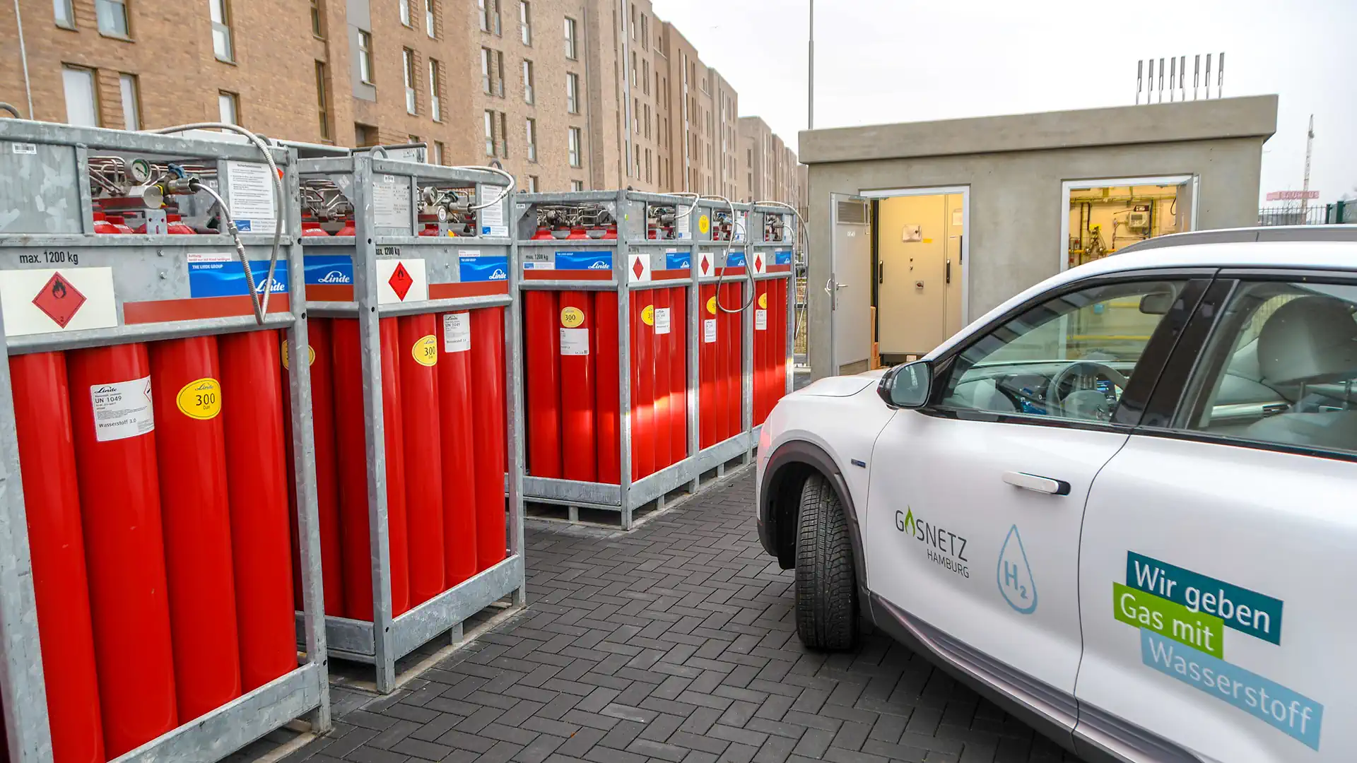 Wasserstoff-Tankstelle mit roten Speicherbehältern und weißem Elektrofahrzeug der Gasnetze Hamburg, Wohngebäude im...