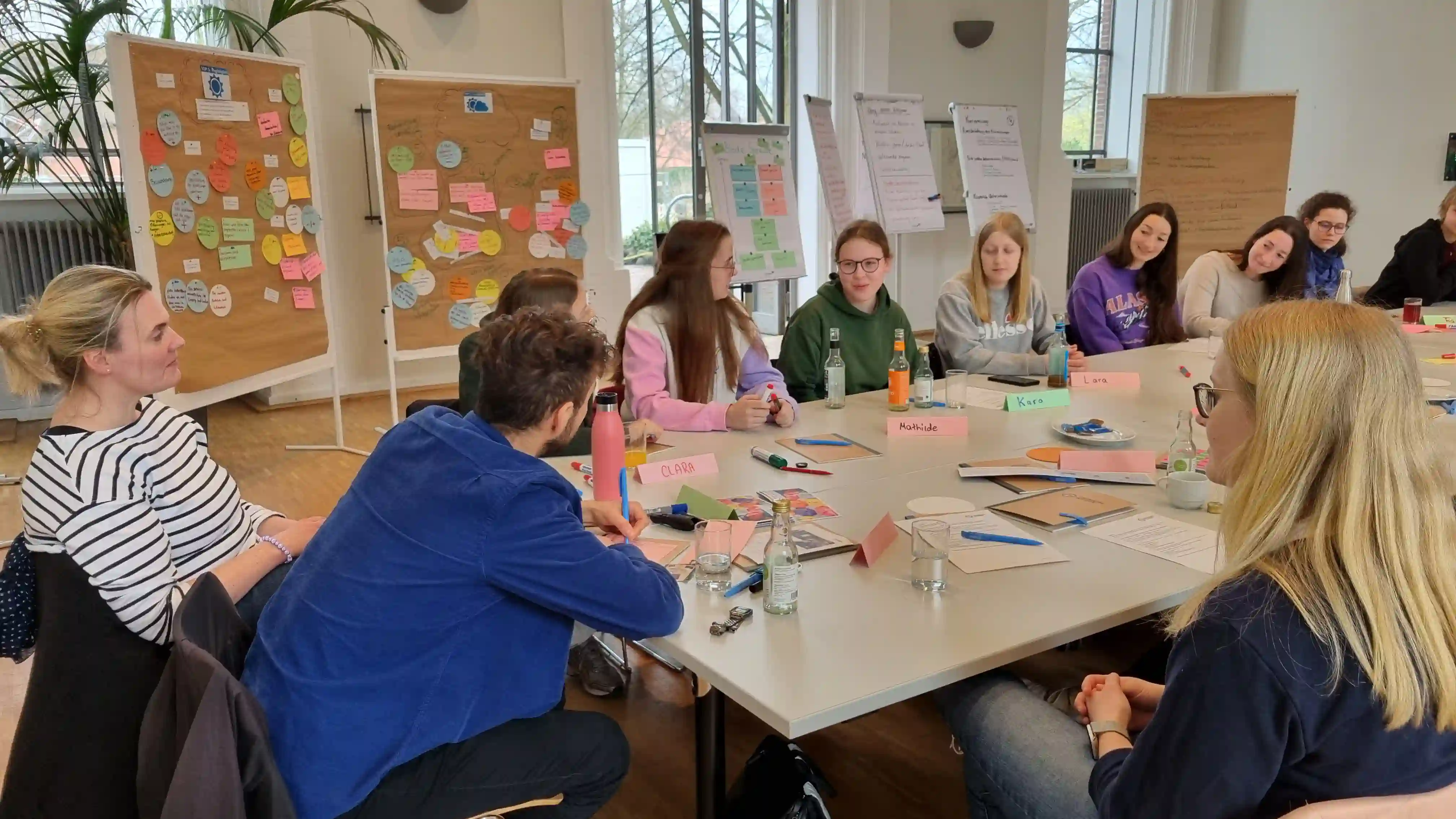 Gruppe von etwa 15 Personen sitzt um Tisch in Workshopraum, dahinter Flipcharts mit bunten Notizen und Korkboards