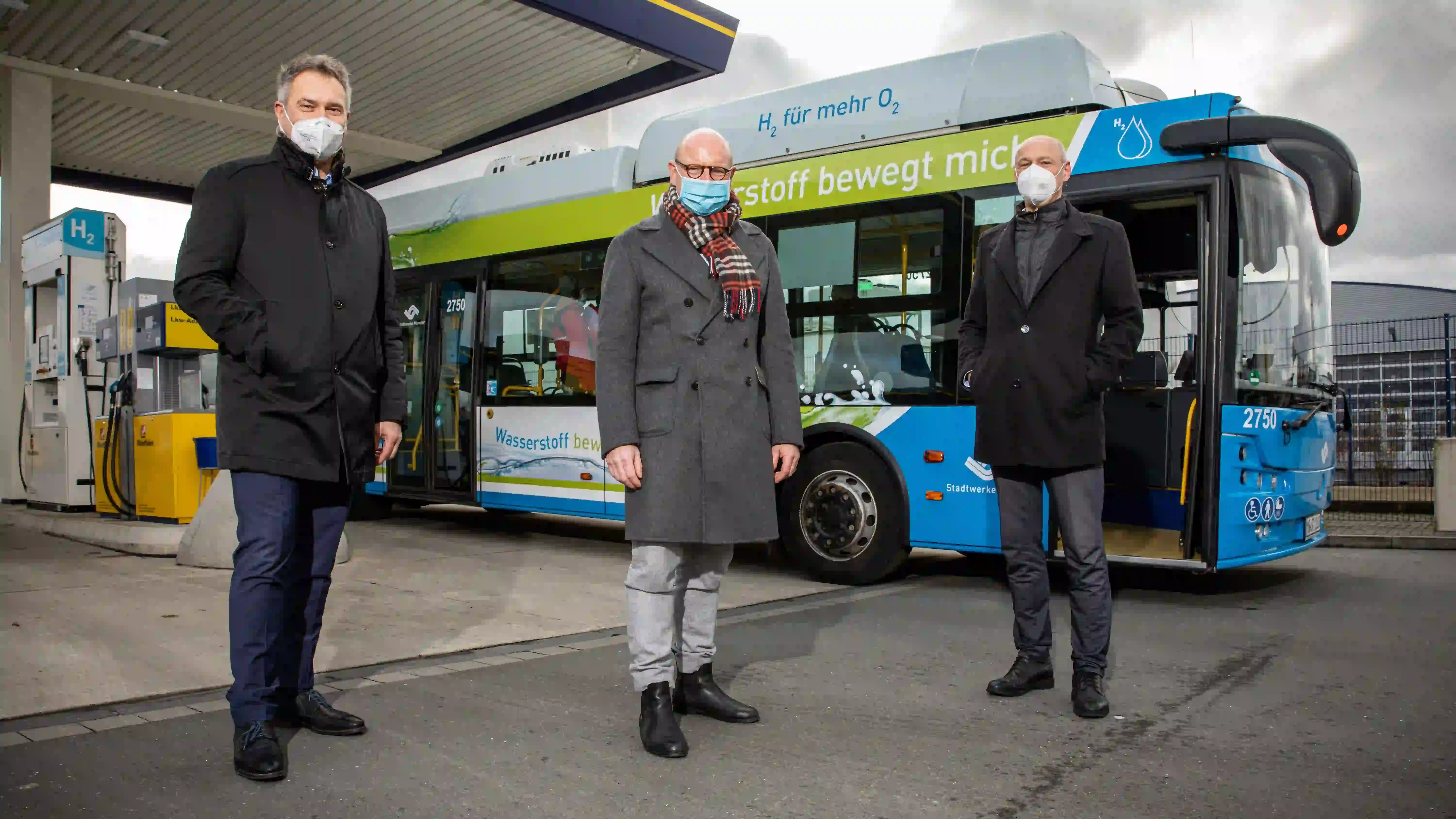 Drei Personen mit Masken stehen vor einem blau-grünen Wasserstoffbus an einer Tankstelle mit H₂-Zapfsäule