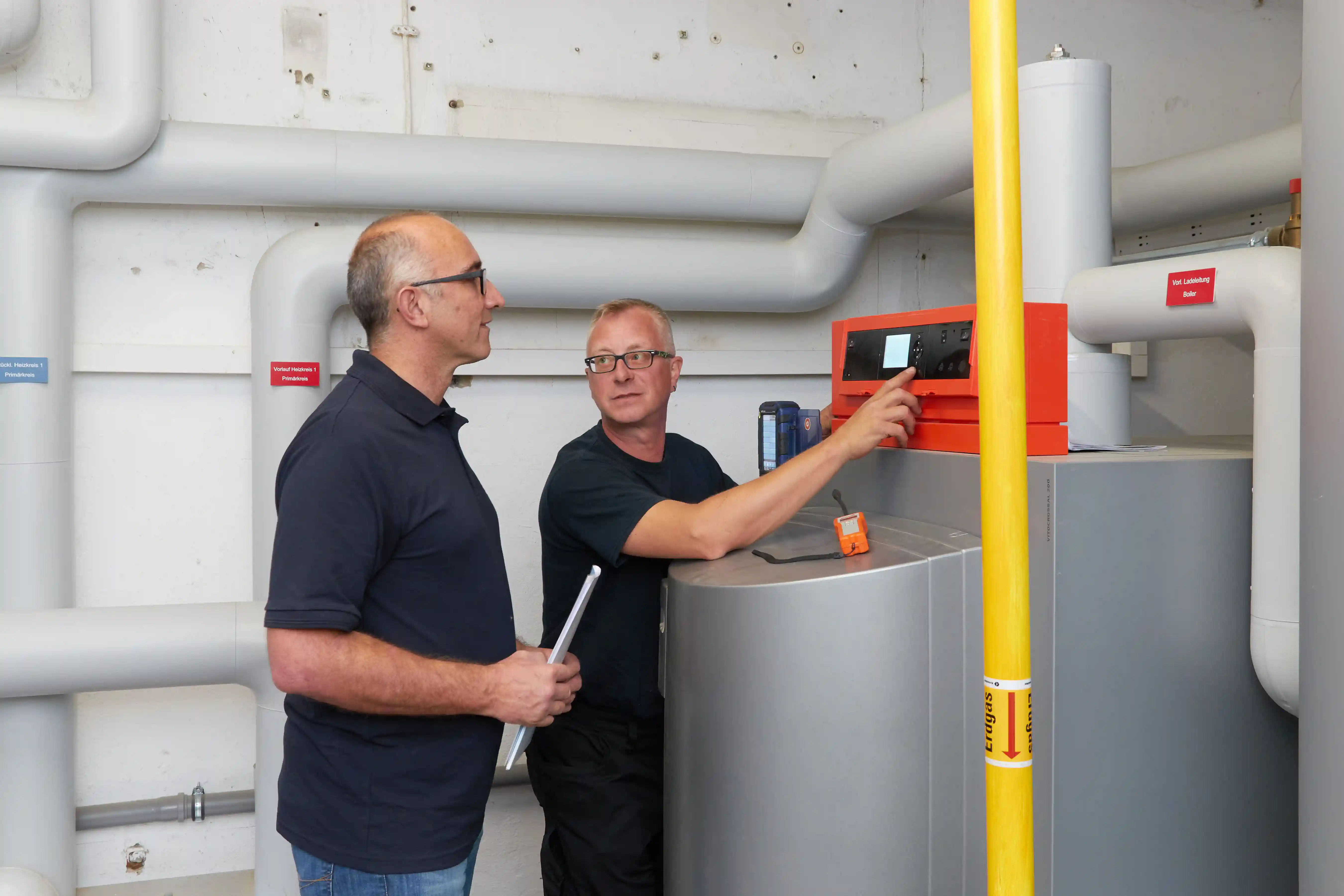 Zwei Techniker inspizieren eine Heizungsanlage mit weißen Rohren und rotem Steuergerät im Heizungskeller.