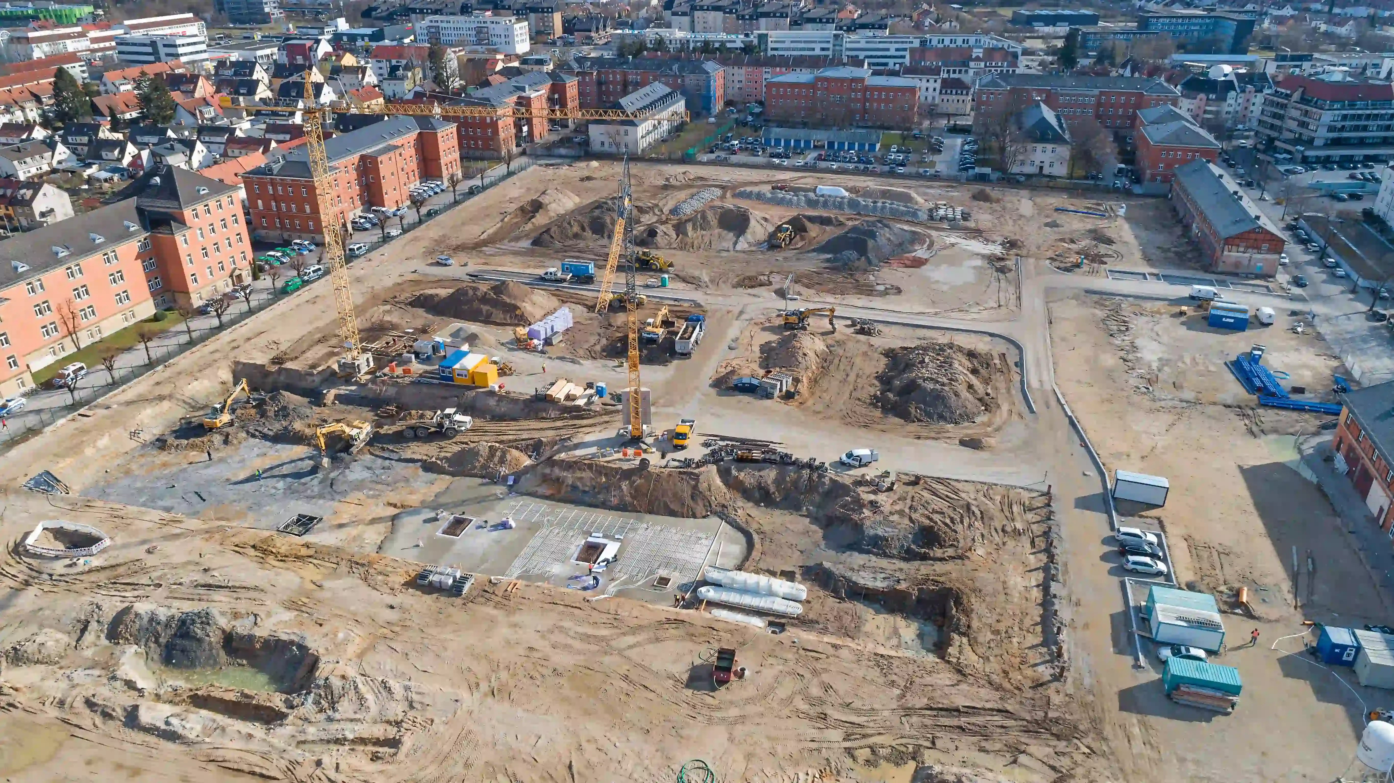 Große Baustelle mit mehreren Kränen und Erdarbeiten in einem urbanen Gebiet mit umliegenden Wohngebäuden.