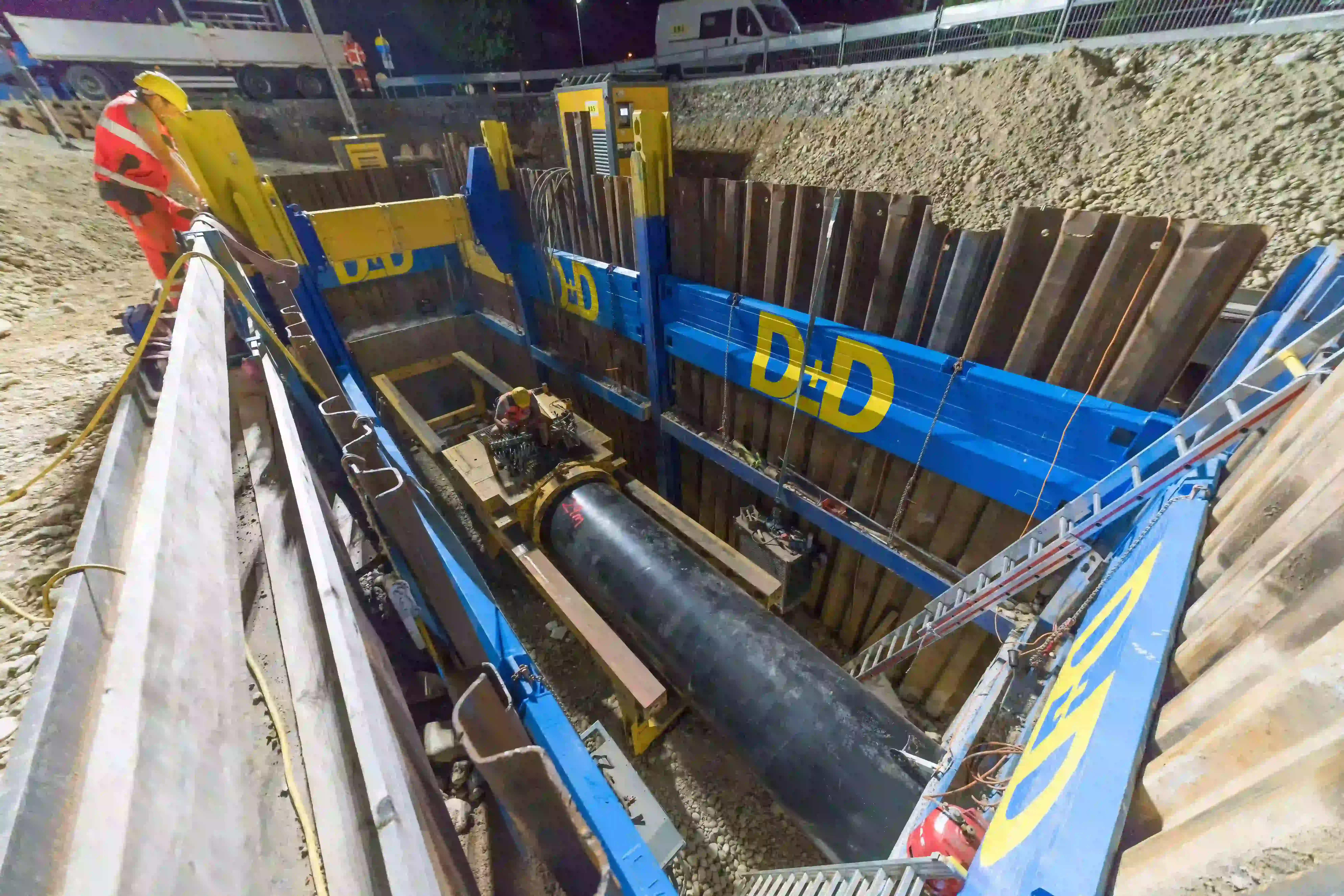 Baugrube mit Stahlverbau und Arbeitern bei der Verlegung einer großen Nahwärme-Rohrleitung bei Nacht.