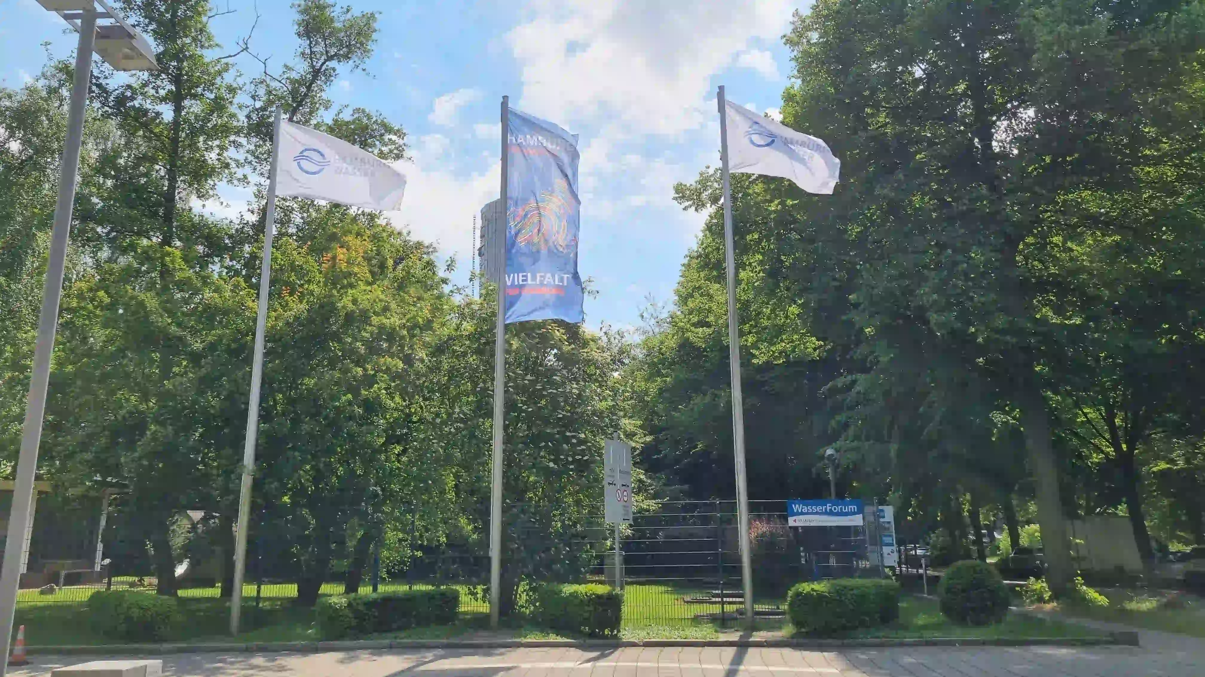 Drei Fahnen vor grünem Baumbestand: zwei weiße Hamburg-Wasser-Flaggen, mittig eine bunte Regenbogenflagge mit...