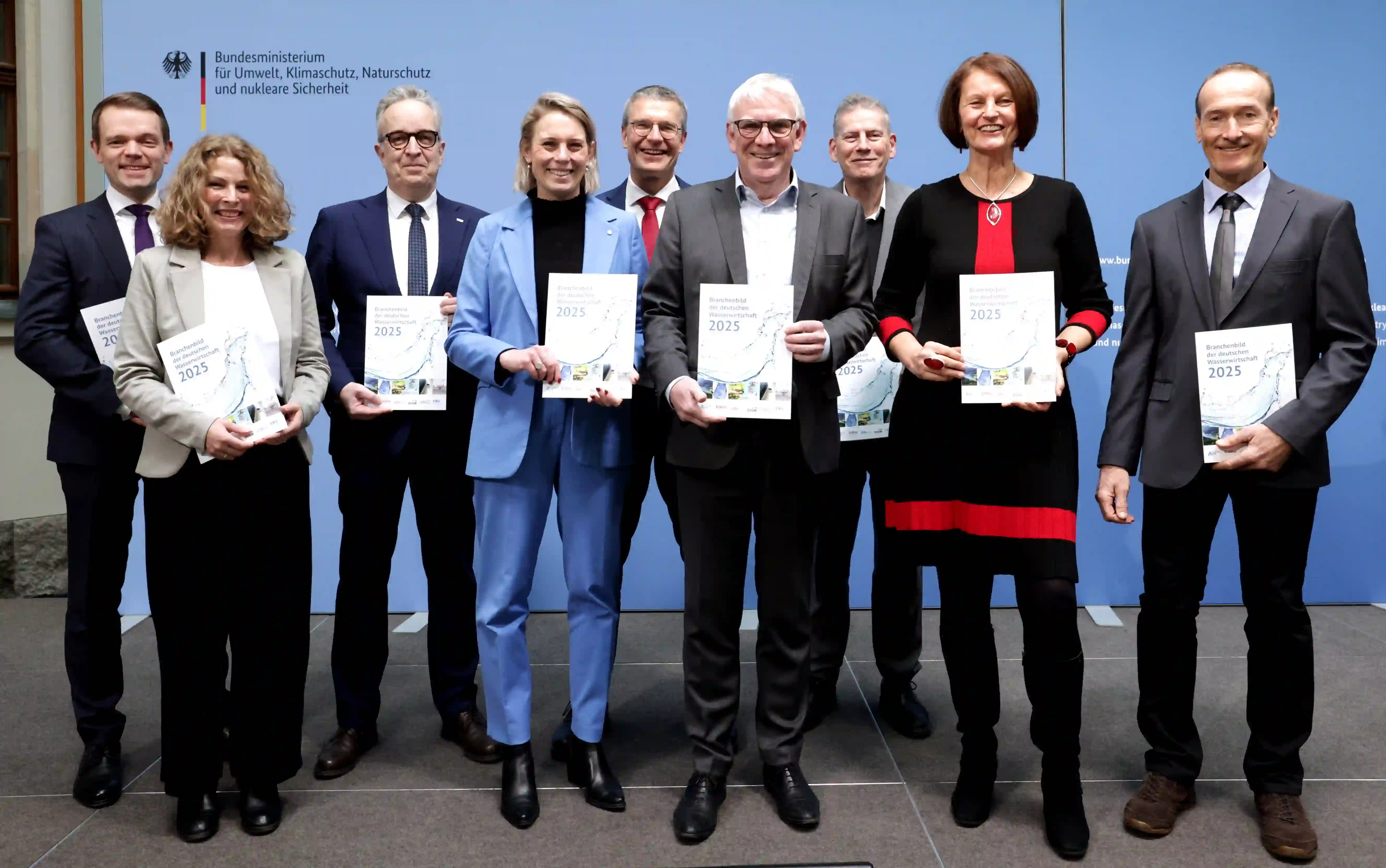 Neun Personen in Business-Kleidung halten Berichte 2025 vor blauem Hintergrund mit Bundesministerium-Logo