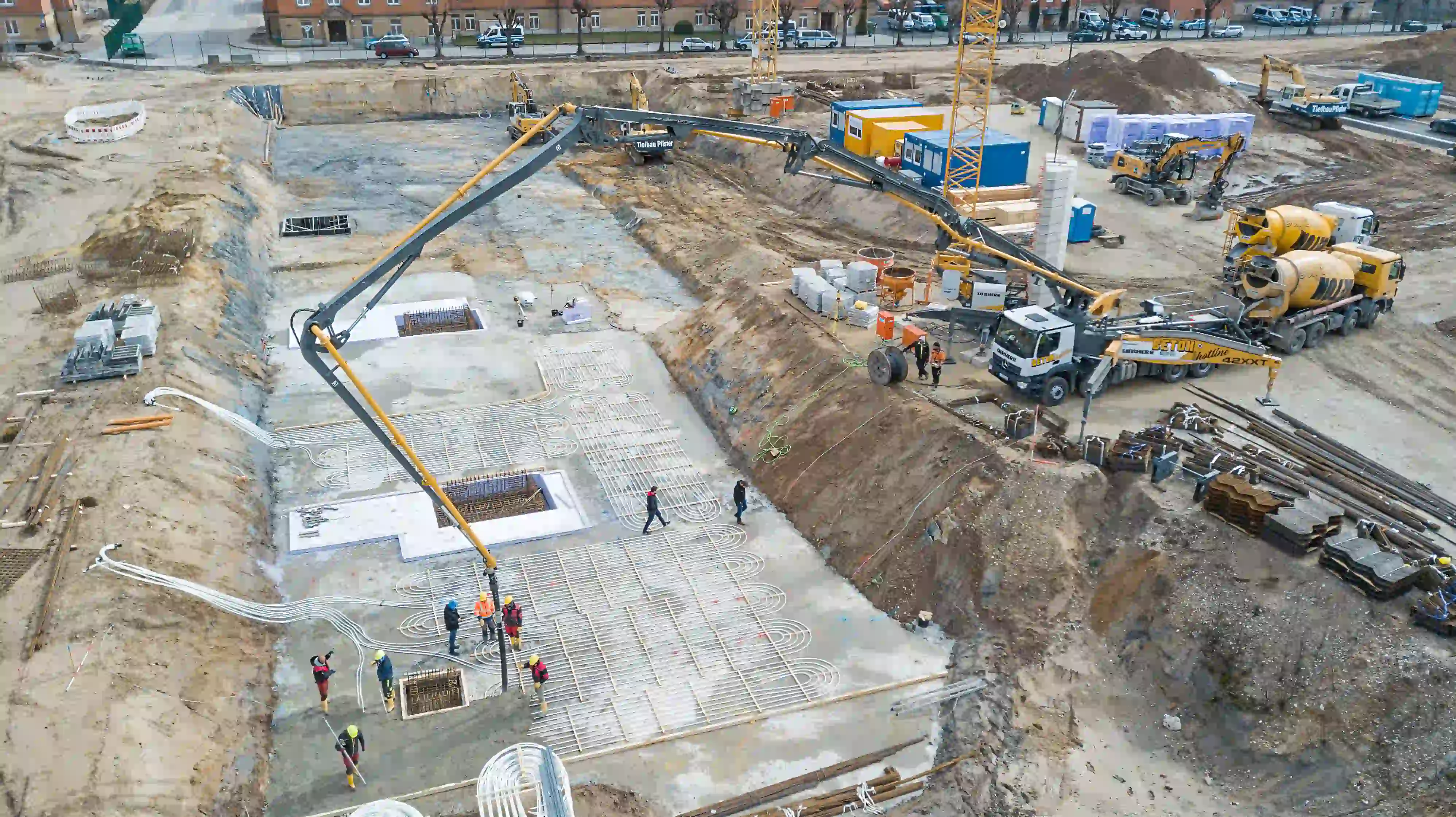 Baustelle mit Erdkollektoren-Verlegung: Arbeiter auf Betonplattform, Bagger und Betonmischfahrzeuge,...