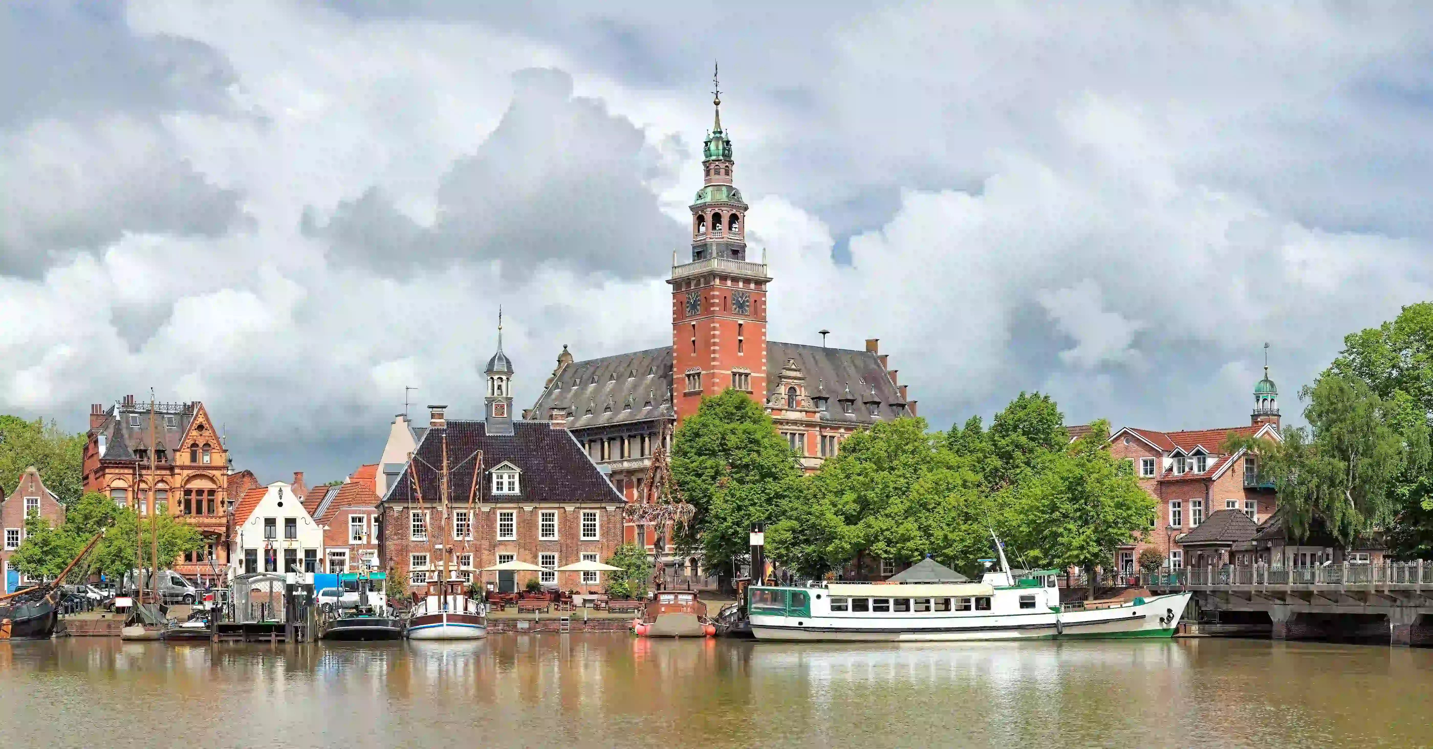 Historische Hafenstadt an einem Fluss mit Backsteingebäuden, rotem Uhrturm und ankernden Booten unter bewölktem Himmel