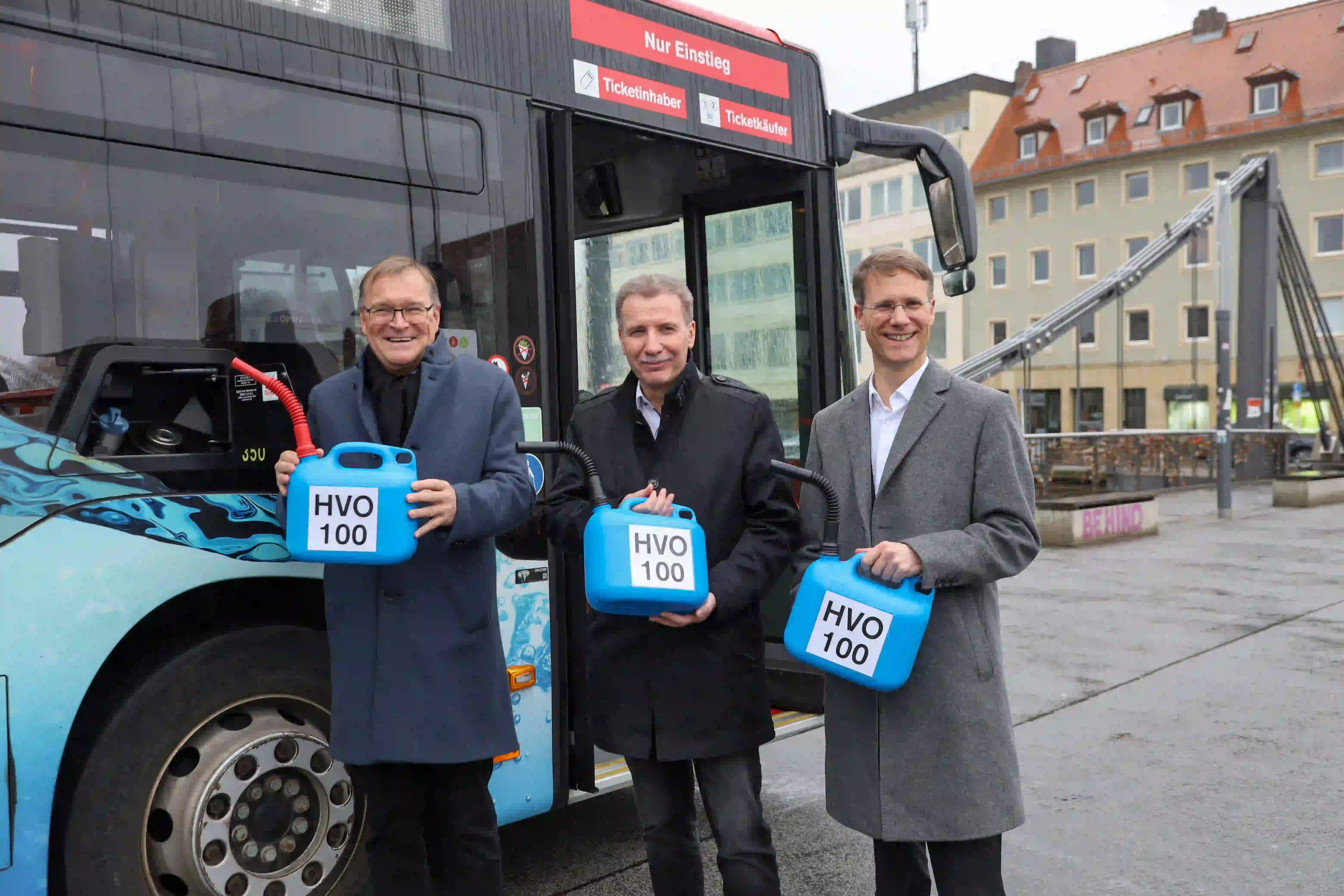 Drei Männer in Mänteln halten blaue Kanister mit der Aufschrift „HVO 100“ vor einem Bus in Bamberg.