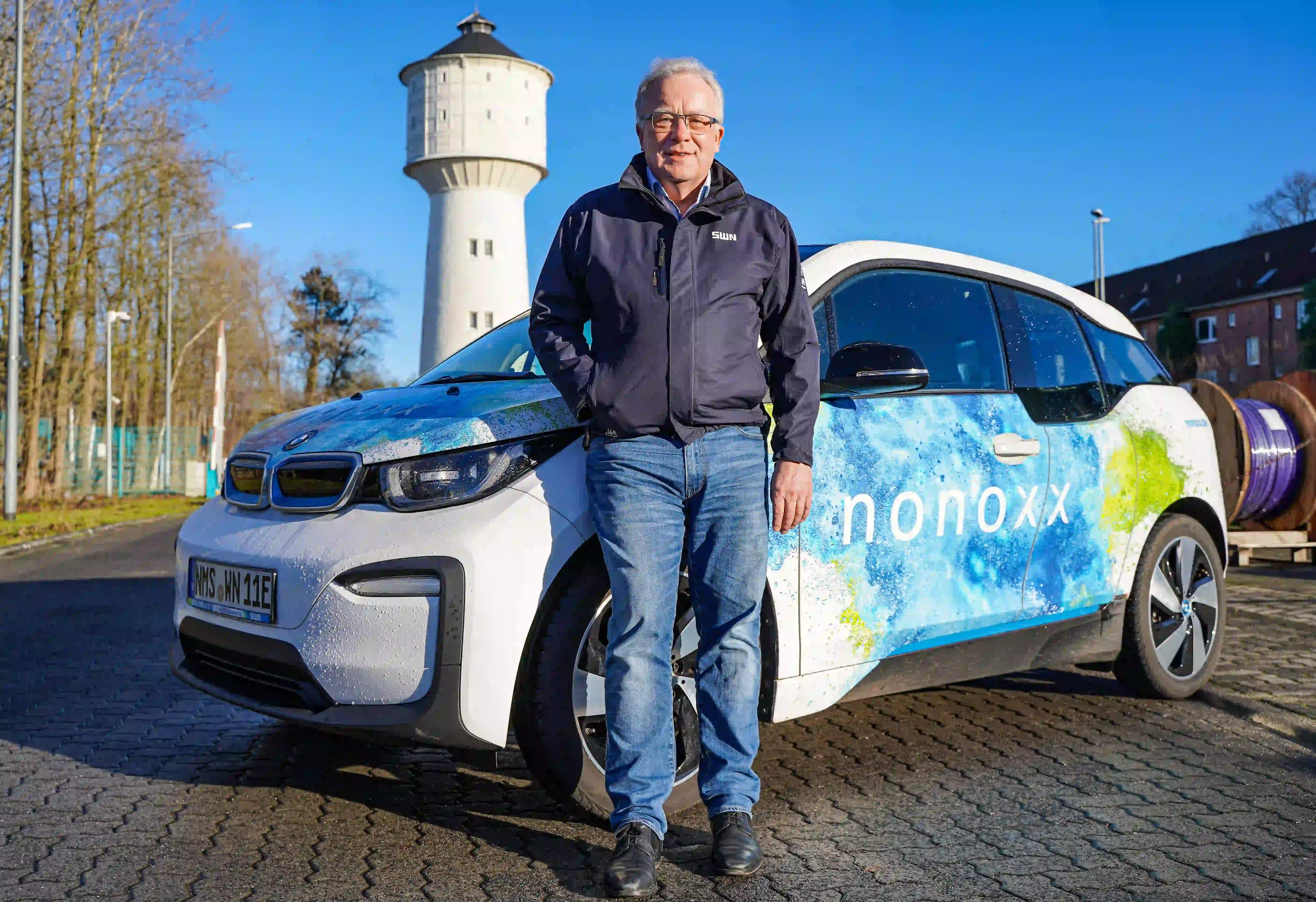 Mann mit Brille steht vor blau-weißem Wasserstoff-Elektroauto, im Hintergrund Wasserturm