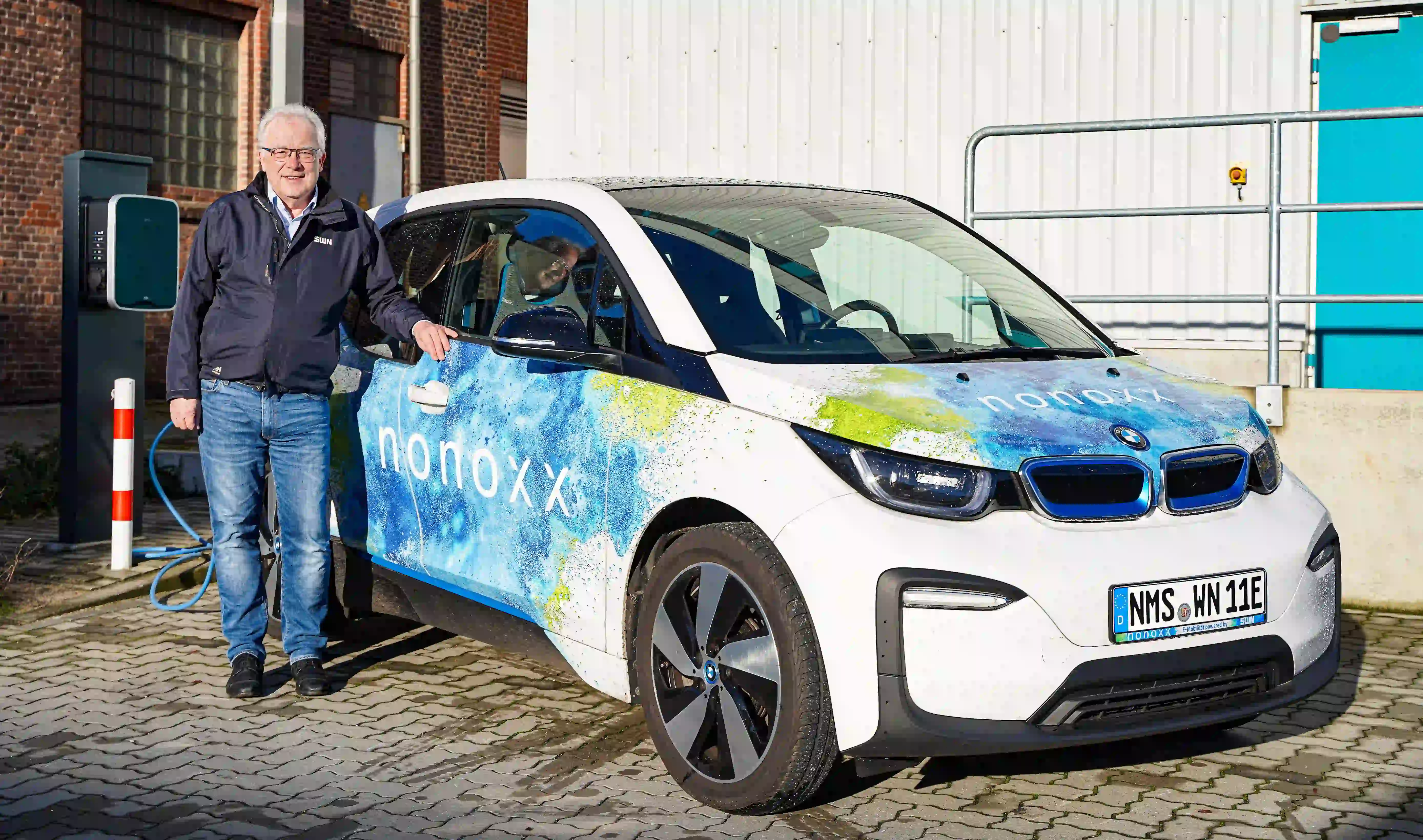 Mann steht an Elektroauto mit Aufschrift „nonoxx“ und lädt es an Ladestation vor Gebäude mit blauer Tür.